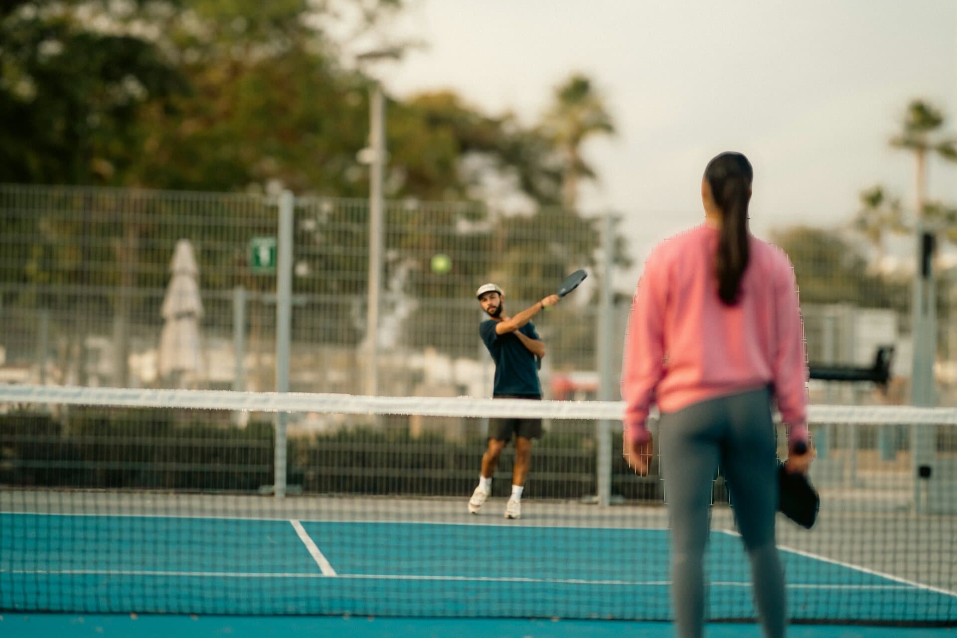 Cuatro personas jugando un partido de dobles de pickleball al aire libre, con la pelota en el aire sobre la red