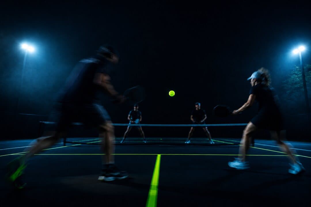 Un rally de dobles de pickleball en una cancha iluminada de noche, con la pelota en el aire sobre la red